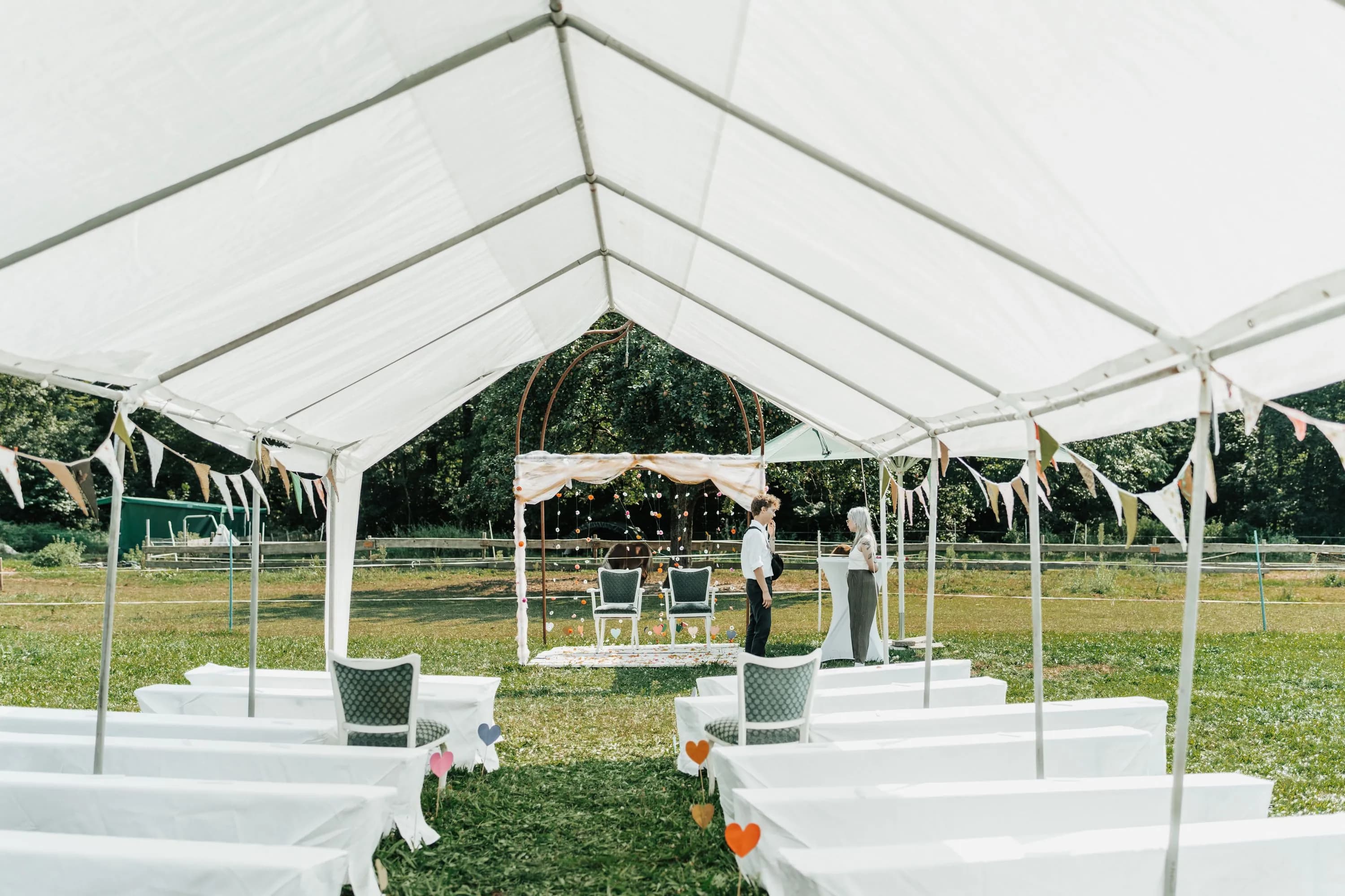 A white frame tent set up with draped tables, chairs, and bunting for an outdoor ceremony